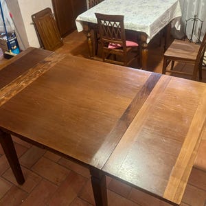 May include: A rectangular wooden dining table with a smooth, medium-brown finish and visible wood grain. The table features a central main surface flanked by two distinct, slightly darker wooden extension panels on either side, supported by four legs.