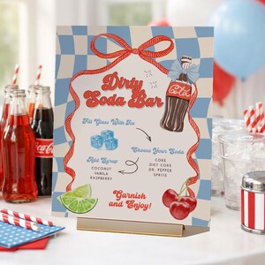 May include: A retro-style "Dirty Soda Bar" sign with a blue and white checkered background. The sign features a Coca-Cola bottle illustration, instructions, and a red ribbon design. It suggests adding syrup and garnishes.