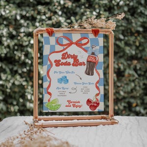 May include: A copper-colored metal frame holds a sign with a blue and white checkered background. The sign reads "Dirty Soda Bar" with illustrations of a soda bottle, ice cubes, and garnishes. The sign includes text for soda flavors and garnishes.