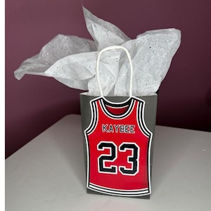 Peut inclure: Un sac cadeau gris avec un motif de maillot de basket-ball rouge. Le maillot porte le nom "KAYBEE" et le numéro "23" en blanc. Le sac est rempli de papier de soie blanc et possède une poignée blanche.