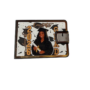 May include: A graduation-themed wallet with a gold-colored frame and clasp. The wallet features a photo of a graduate in a cap and gown, with the words "Congrats Grad" and "Class of 2026" printed on the front.