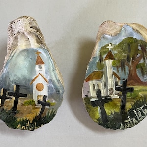 May include: Two oyster shells painted with church scenes. Each shell features a white church with a brown steeple and crosses in the foreground. The backgrounds have a blue sky and green grass. The artist's signature is visible on one shell.