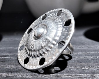 May include: A silver ring with a large, round, decorative top. The top has a raised, circular design with a smaller, round, raised center piece. The ring has several small holes around the edge of the top.