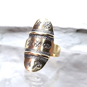Puede incluir: Un anillo dorado con una cara ovalada alargada. El anillo presenta intrincados patrones y diseños grabados, que incluyen formas geométricas y líneas. La banda es de un color dorado liso y sencillo.