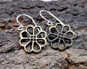 May include: A pair of flower-shaped earrings. Each earring features a gold-colored, openwork floral design with a silver-colored hook. The earrings are displayed on a textured, dark brown surface.