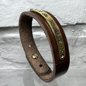 May include: Brown leather bracelet with a gold-colored metal plate. The plate has etched symbols. The bracelet has a buckle closure with a gold-colored stud. The leather is a medium brown color.