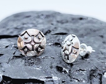 May include: A pair of silver stud earrings with a circular design featuring a geometric pattern. The earrings have a textured surface and a slightly raised center.