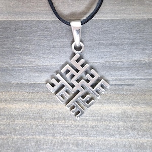 Peut inclure: Un pendentif argenté avec un motif géométrique complexe, suspendu à un cordon noir. Le pendentif est de forme carrée, avec un motif complexe de lignes et de formes interconnectées. Le fond est une surface en bois grise.