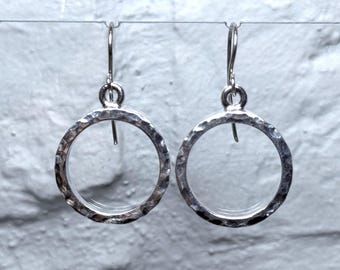 May include: A pair of silver-colored hoop earrings with a hammered texture. The earrings are circular with a simple hook closure. The background is a white brick wall.