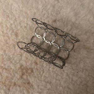 May include: A silver-colored bracelet composed of interconnected, hammered metal rings. The bracelet is flexible and appears to be made of a lightweight metal. The design is open and airy, with a geometric pattern. The bracelet is shown on a textured, off-white surface.