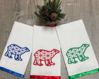 May include: Three white kitchen towels with a waffle weave texture. Each towel features a different color embroidered polar bear with a snowflake pattern. The bears are blue, red, and green.
