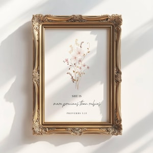 May include: A framed print with a gold ornate frame. The print features a floral arrangement in soft pink and beige hues. The text reads "SHE IS more precious than rubies" from Proverbs 3:15.