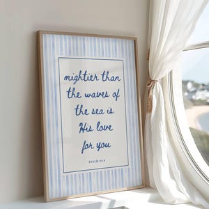 Op de afbeelding: Een ingelijste print met de tekst "mightier than the waves of the sea is His love for you" in blauw schrift. De print heeft een lichtblauwe en witte gestreepte achtergrond en een houten lijst. De print staat op een vensterbank naast een raam met uitzicht op de zee.