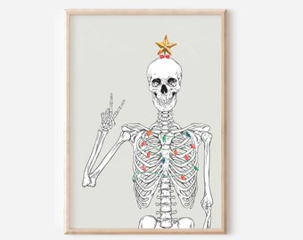 Op de afbeelding: Een ingelijste print met een skelet versierd met een gouden ster en kerstverlichting. Het skelet maakt het vredesteken. Het kunstwerk heeft een lichtgrijze achtergrond en wordt gepresenteerd in een houten lijst.