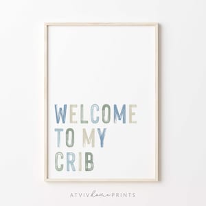 May include: A framed nursery print with the text "Welcome to my crib" in blue, green, and beige brushstrokes. The print is on a white background and has the text "ATVIV home PRINTS" at the bottom.