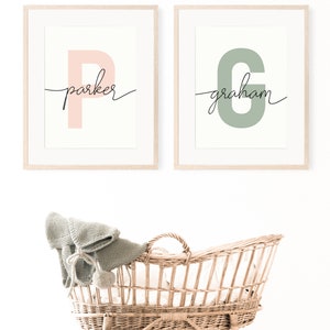 May include: Two framed prints with the letters "P" and "G" in pink and green, respectively. The letters are stylized and have the names "parker" and "graham" written in cursive underneath. A wicker bassinet with a knitted blanket sits in front of the prints.