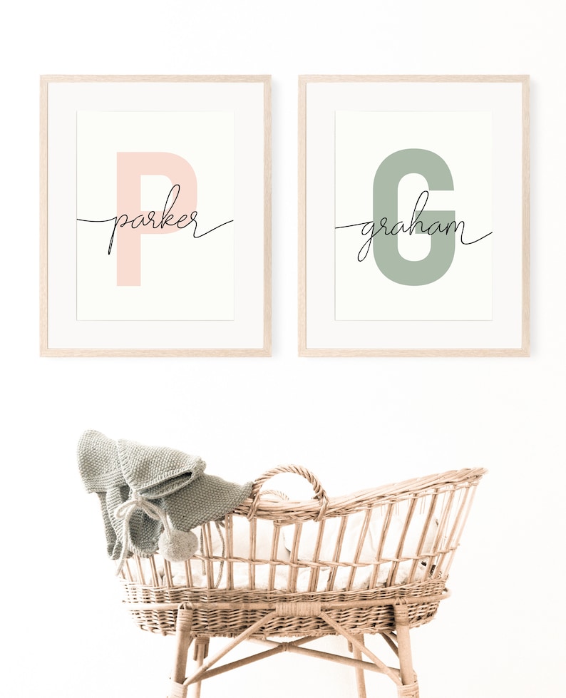 May include: Two framed prints with the letters "P" and "G" in pink and green, respectively. The letters are stylized and have the names "parker" and "graham" written in cursive underneath. A wicker bassinet with a knitted blanket sits in front of the prints.