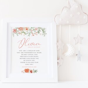 Puede incluir: Una impresi&oacute;n enmarcada en blanco con detalles florales de acuarela en melocot&oacute;n y verde, con el nombre Olivia Grace Watson y texto religioso en letras negras y melocot&oacute;n, incluyendo la fecha Baptized March 28, 2022, junto a un m&oacute;vil de nube de peluche.