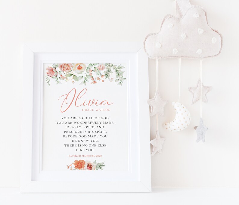 Puede incluir: Una impresi&oacute;n enmarcada en blanco con detalles florales de acuarela en melocot&oacute;n y verde, con el nombre Olivia Grace Watson y texto religioso en letras negras y melocot&oacute;n, incluyendo la fecha Baptized March 28, 2022, junto a un m&oacute;vil de nube de peluche.