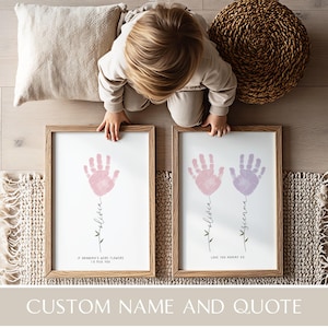 Può includere: Due stampe incorniciate con arte di impronte di mani. Una stampa presenta un'impronta rosa con il nome "Olivia" e la citazione "If Grandma's were flowers I'd pick you." L'altra stampa ha un'impronta viola con il nome "Sienna" e la citazione "Love you Mommy xx."