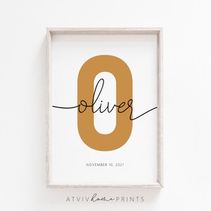 May include: A printable art print with a gold number zero and the name "Oliver" written in cursive script. The date "November 10, 2021" is printed below the name. The print is framed in a light wood frame.