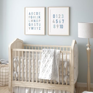 May include: A white wooden crib with a white quilted blanket and a white ruffle skirt. Two framed prints hang on the wall behind the crib. The print on the left shows the alphabet in blue and gray. The print on the right shows the numbers 0-10 in blue and gray.
