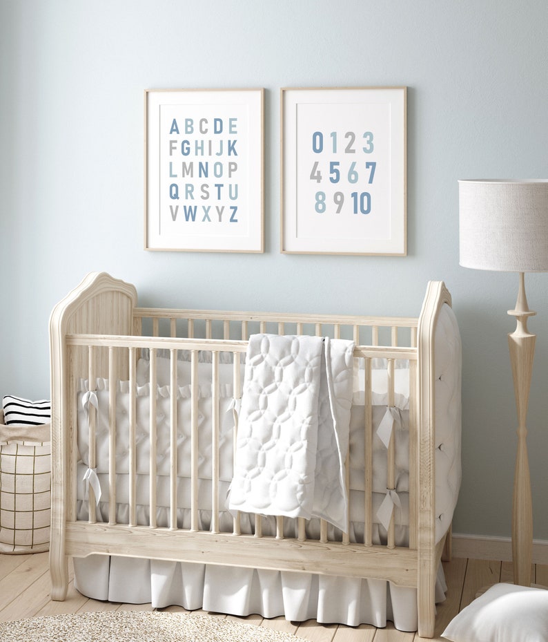 May include: A white wooden crib with a white quilted blanket and a white ruffle skirt. Two framed prints hang on the wall behind the crib. The print on the left shows the alphabet in blue and gray. The print on the right shows the numbers 0-10 in blue and gray.