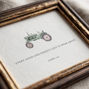 Op de afbeelding: Een ingelijste print met een aquarelillustratie van een groene tractor. Onder de tractor staat de tekst: EVERY GOOD AND PERFECT GIFT IS FROM ABOVE JAMES 1:17. Het kunstwerk zit in een donkerbruine houten lijst met gouden accenten.