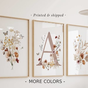 Może przedstawiać: Trzy oprawione botaniczne wydruki artystyczne. Wydruki przedstawiają akwarelowe ilustracje kwiatów, motyli i inicjału. Ramki są z jasnego drewna. Widoczny jest tekst "Printed & shipped" i "MORE COLORS".