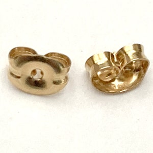 May include: Two gold-colored earring backs. The backs have a butterfly shape with a central hole for the earring post. The metal has a shiny finish. These are small jewelry components.