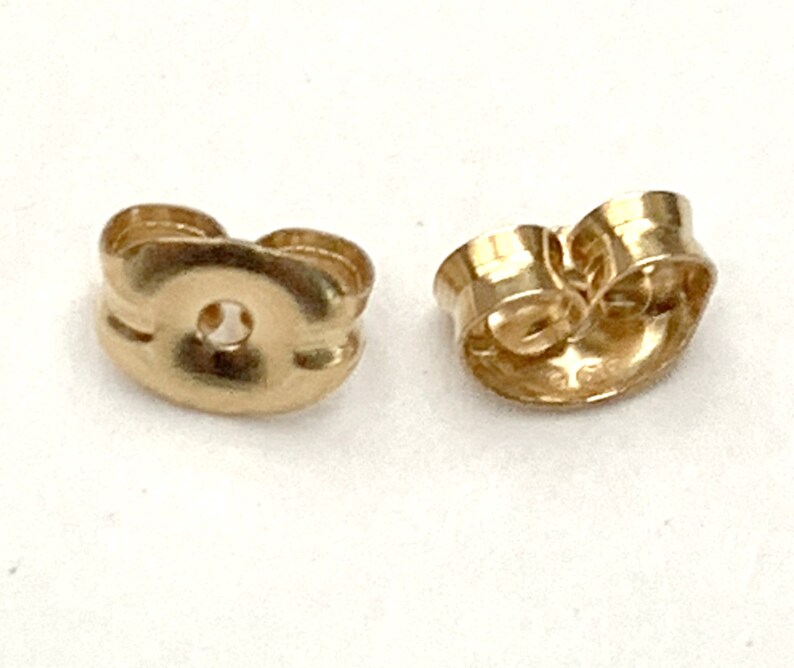 May include: Two gold-colored earring backs with a rounded, butterfly-style shape. Each piece features a central circular opening designed to secure an earring post, with curved metal wings extending outward to provide a firm grip for the wearer.