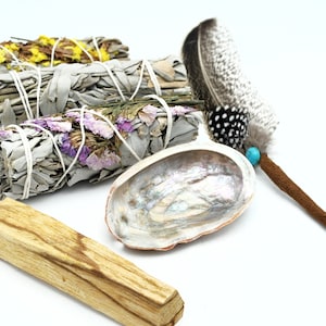 May include: A set of smudging tools including white sage bundles with dried flowers, a palo santo stick, a seashell, and a feather with a turquoise bead.