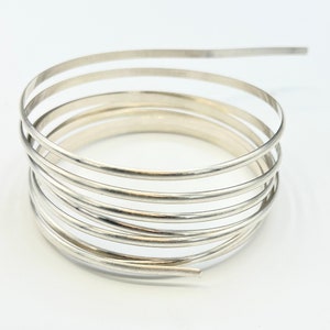May include: A silver-colored, coiled bracelet. The bracelet is made of multiple, thin, rounded bands that spiral around each other. The design is simple and modern, with a polished finish. The bracelet is shown against a white background.