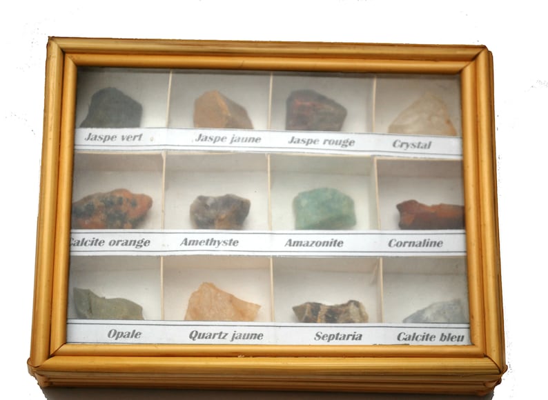 May include: A wooden display case with 12 compartments, each containing a different type of rock or mineral. The case has a glass front and is labeled with the names of the rocks in French. The rocks include jaspe vert, jaspe jaune, jaspe rouge, crystal, calcite orange, amethyste, amazonite, cornaline, opale, quartz jaune, septaria, and calcite bleu.