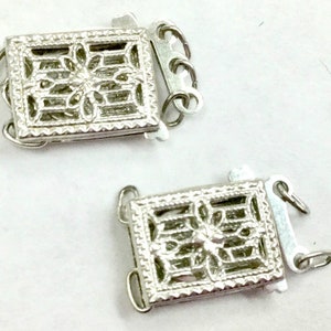 May include: Two silver-toned metal clasps with intricate filigree designs. Each clasp has a rectangular shape with a decorative floral pattern and a hook closure.