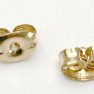 May include: Two gold-colored metal earring backs with a butterfly clutch design. Each piece features a curved, rounded shape with a central hole for an earring post and two small, coiled metal loops that provide tension to secure the earring.