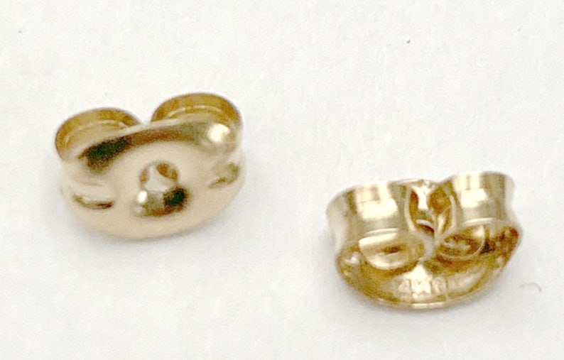 May include: Two gold-colored metal earring backs with a butterfly clutch design. Each piece features a curved, rounded shape with a central hole for an earring post and two small, coiled metal loops that provide tension to secure the earring.