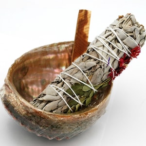 May include: A white sage smudge stick with dried flowers and herbs, tied with white string, resting in a brown abalone shell.