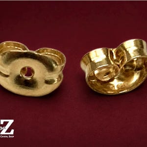 May include: Two gold-colored metal earring backs with a curved, butterfly-shaped design. Each piece features a central circular opening for an earring post and a smooth, reflective surface. The logo A-Z Jewelry Supply & Crystal Shop is visible.