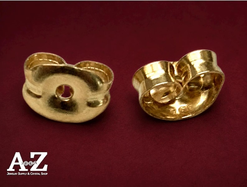 May include: Two gold-colored metal earring backs with a curved, butterfly-shaped design. Each piece features a central circular opening for an earring post and a smooth, reflective surface. The logo A-Z Jewelry Supply & Crystal Shop is visible.
