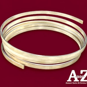 Puede incluir: Una longitud enrollada de alambre metálico plano de color dorado dispuesto en forma de espiral. El alambre tiene una superficie lisa y reflectante con una sección transversal rectangular constante, formando múltiples bucles concéntricos.