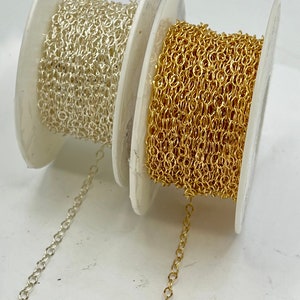 May include: Two spools of chain, one gold and one silver, are shown on a white surface. The chain is unraveled and hanging down from the spools.