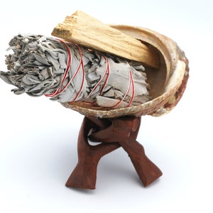 Puede incluir: Un manojo de salvia blanca atado con cuerda roja y un trozo de palo santo, descansando en una concha de mar sobre un soporte de madera.