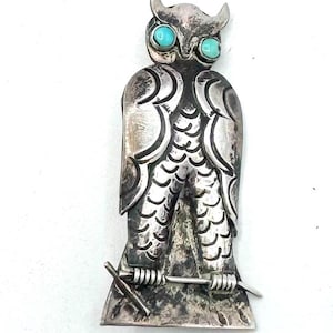 May include: A silver-toned owl-shaped brooch with turquoise-colored eyes. The brooch features detailed feather and wing patterns, and a small, stylized branch at the bottom. The owl has small horns and is set against a white background.