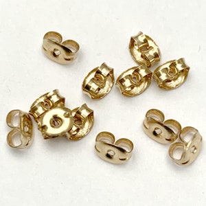 May include: A collection of eight gold-toned earring backs, each featuring a small circular center hole and a curved, butterfly-shaped metal design. These small jewelry components are scattered across the frame, showing their metallic finish.