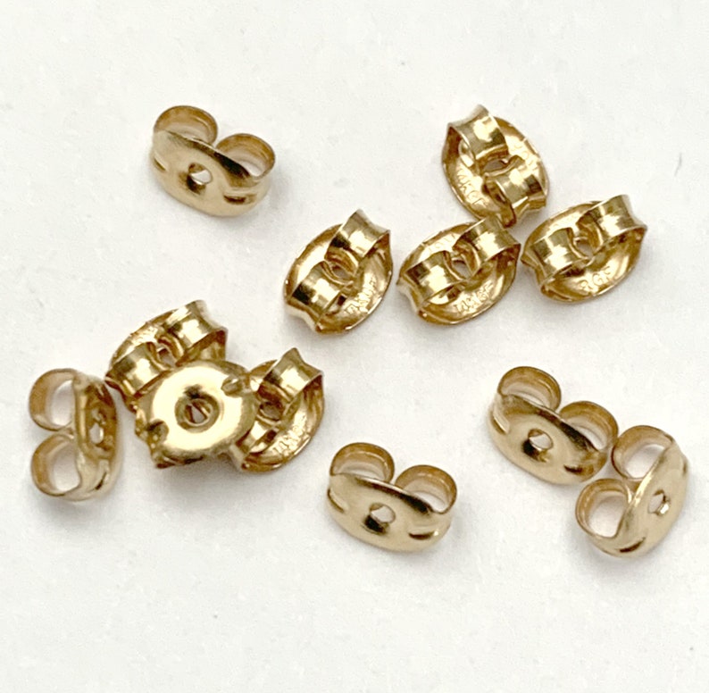 May include: A collection of eight gold-toned earring backs, each featuring a small circular center hole and a curved, butterfly-shaped metal design. These small jewelry components are scattered across the frame, showing their metallic finish.