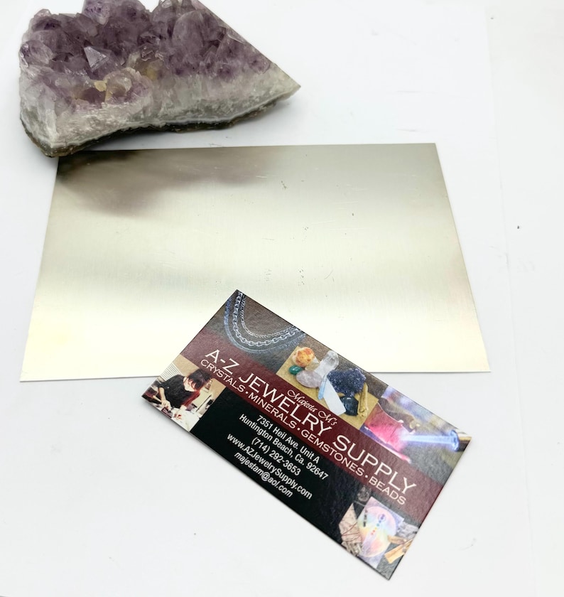 Puede incluir: Una tarjeta de visita para A-Z Jewelry Supply, con un fondo rojo y texto blanco. La tarjeta incluye el nombre de la empresa, la direcci&oacute;n, el n&uacute;mero de tel&eacute;fono, el sitio web y la direcci&oacute;n de correo electr&oacute;nico. La tarjeta tambi&eacute;n incluye una peque&ntilde;a imagen de una variedad de cristales y piedras preciosas.