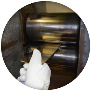 Puede incluir: Una l&aacute;mina de metal est&aacute; siendo enrollada por una m&aacute;quina con dos grandes rodillos de metal. Una mano enguantada est&aacute; guiando la l&aacute;mina de metal a trav&eacute;s de los rodillos.