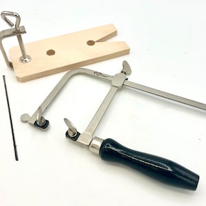 May include: A silver-toned jewelry saw with a black handle and a wooden saw frame with a clamp. The saw is used for cutting metal jewelry.