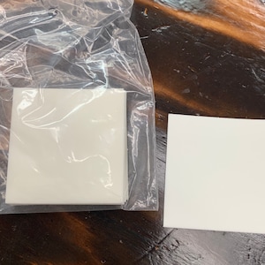 May include: A stack of white square paper sheets in a clear plastic bag, and a single white square paper sheet on a wooden surface.